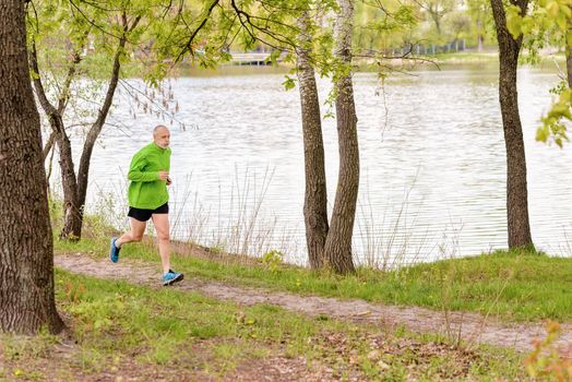 A tired senior man dressed in black and green is running in the park, close to the lake, during a gray spring day