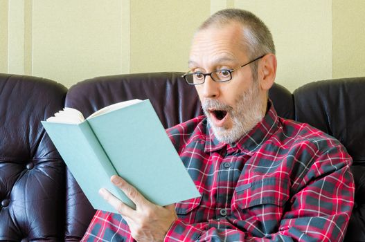 A surprised senior man with beard and spectacles, sitting on the sofa and reading a book