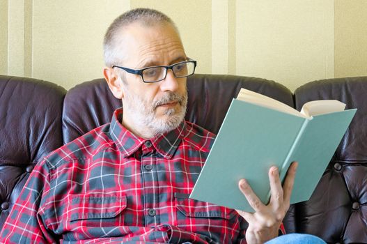 A senior man with beard and spectacles, sitting on the sofa and reading a book