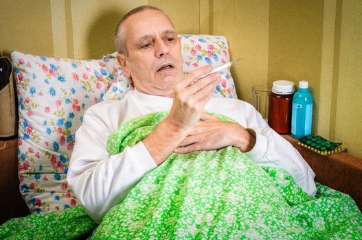 Sick man lying in bed and measuring the temperature with a thermometer