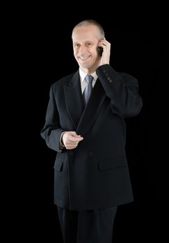 An agreeable businessman wearing a black suit smiling while speaking on mobile phone, on black background