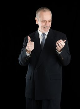 An amiable businessman wearing a black suit  smiling and reading on mobile phone, with thumb up, on black background