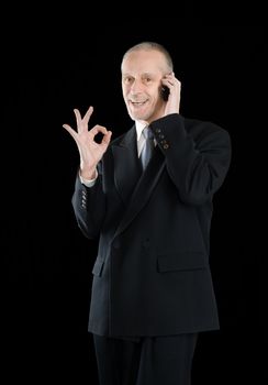 An amiable businessman in black suit, smiling on mobile phone and showing the okay sign with the hand, on black background