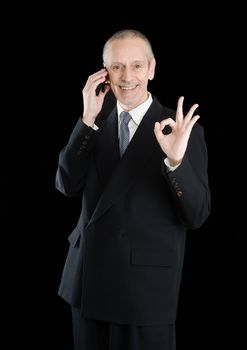 An amiable businessman in black suit, smiling on mobile phone and showing the okay sign with the hand, on black background