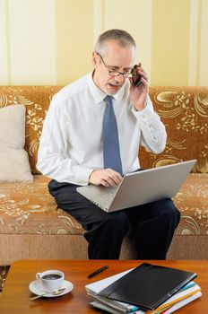 An handsome senior businessman working with computer at home, calling to a partner with his mobile phone and ready to drink a hot cup of black coffee
