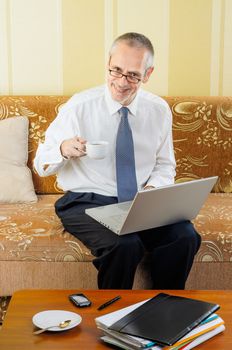 An handsome senior businessman working with computer at home, holding spectacles in his hand and ready to drink a hot cup of black coffee