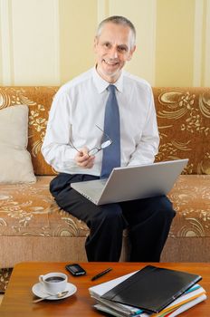 An handsome senior businessman working with computer at home, holding spectacles in his hand and ready to drink a hot cup of black coffee