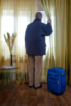 A man with a blue suitcase, ready to start for a travel, moves the curtain aside to look trough the window  if the taxi is coming