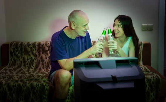 An adult man and woman watch sports on television in their small apartment. Their favorite team won the game.  They drink to his victory, smiling full of joy and happiness