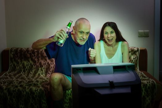 An adult man and woman watch sports on television. Their favorite team scored a goal and they react as good supporters, expressing their happiness with laughter and howl of joy