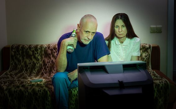 Middle-aged man and woman, watch television, looking sad, sitting on a couch. They watch a sad movie or listen to bad news that makes them a little unhappy and depressed