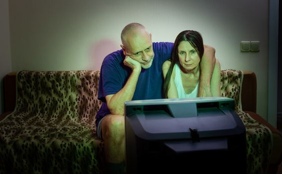 A couple of adult lovers, sitting on a couch covered with a leopard pattern blanket, watch a romantic movie on television. There is a little tenderness and nostalgia in their eyes, a little sadness