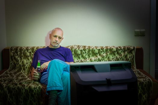 An adult man, alone, watches television, looking sad, sitting on a couch, with a bottle of beer in the hand. He watches a sad movie or listens to bad news that makes him a little unhappy and depressed