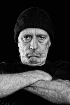 Dramatic black and white photo of a strong man with a mutton chops beard with a woolen cap on the head, crossing the arms with an agressive look.