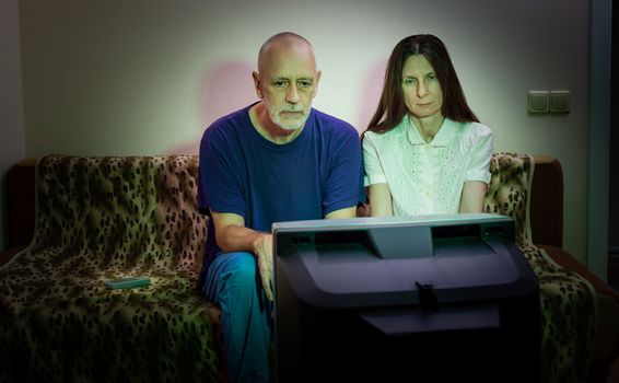 Adult man and woman, watch television, looking sad, sitting on a couch. They watch a sad movie or listen to bad news that makes them a little unhappy and depressed