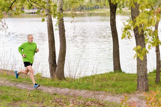 A senior man dressed in black and green is running in the park, close to the lake, during a gray spring day