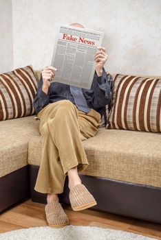 A man wearing glasses is sitting on a couch at home, reading a newspaper reporting fake news. Fake Lorem ipsum text.