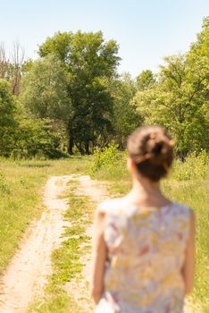 A woman with a chignon, standing up close to the country road in Kiev, Ukraine, observes the trees in the distance. The silhouette of the lady is out of focus, against a focused background.