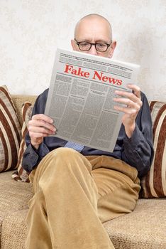 A man wearing glasses is sitting on a couch at home, reading a newspaper reporting fake news. Fake Lorem ipsum text.