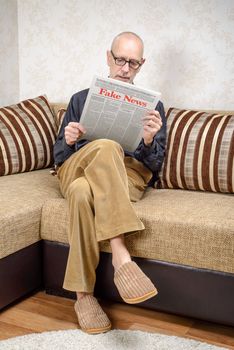 A man wearing glasses is sitting on a couch at home, reading a newspaper reporting fake news. Fake Lorem ipsum text.