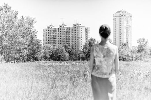 Unfocussed woman looking at modern residential buildings while see stands in the country, in the middle of the meadows, under the sun during a warm spring day