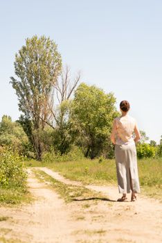 A woman with a chignon, standing up close to the country road in Kiev, Ukraine, observes the trees in the distance. The silhouette of the lady is out of focus, against a focused background.