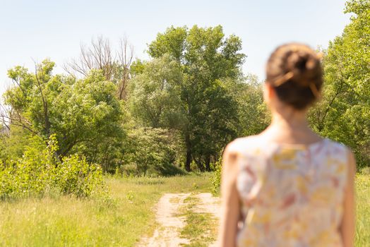 A woman with a chignon, standing up close to the country road in Kiev, Ukraine, observes the trees in the distance. The silhouette of the lady is out of focus, against a focused background.