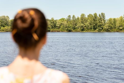 A woman with a chignon, standing up close to the Dnieper river in Kiev, Ukraine, observes the trees in the distance. The silhouette of the lady is out of focus, against a focused background.