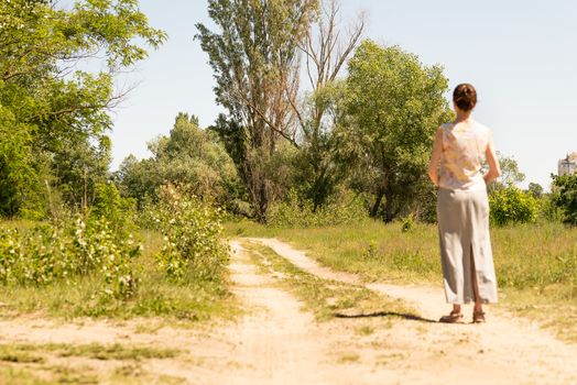 A woman with a chignon, standing up close to the country road in Kiev, Ukraine, observes the trees in the distance. The silhouette of the lady is out of focus, against a focused background.