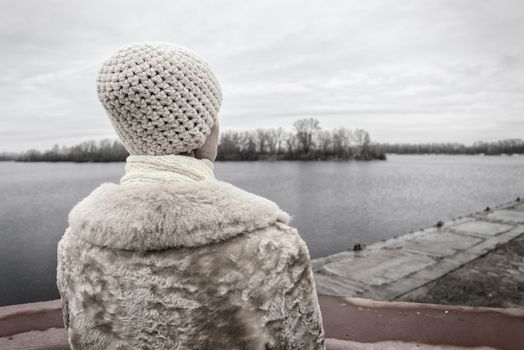 A woman with a wool cap and a fur coat is looking at the river during a cold and sad gray winter morning