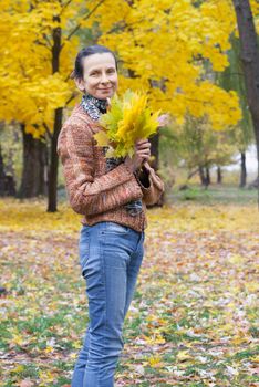 A smiling adult caucasian woman is picking yellow maple leaves in autumn in the park