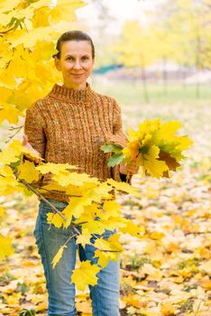 A smiling adult caucasian woman is picking yellow maple leaves in autumn in the park