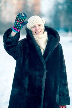 A winter portrait of a smiling senior adult woman wearing a wool cap, a scarf and colored gloves, saluting with her hand, with a snow background