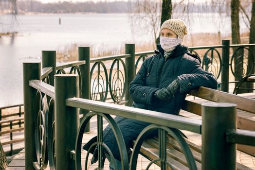 A poor elderly woman wears a homemade mask to protect herself from viruses such as coronavirus, also known as covid-19, or SARS and MERS. She sits on a bench close to the river