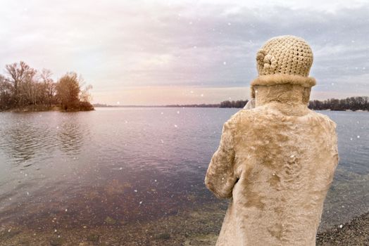 A woman with a wool cap and a fur coat is looking at the river during a cold and sad gray winter morning under the snow