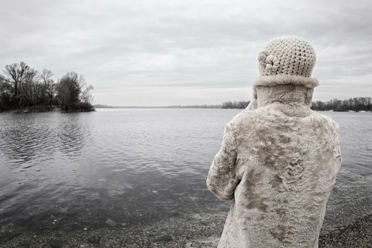 A woman with a wool cap and a fur coat is looking at the river during a cold and sad gray winter morning