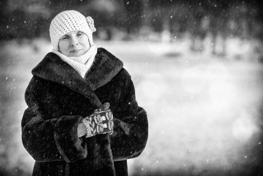 A winter portrait of a smiling senior adult woman wearing a wool cap, a scarf and colored gloves, with a snow background