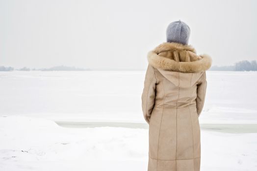 A woman with a wool cap and a leather coat is looking at the Dnieper river during a cold and sad gray winter morning under the snow