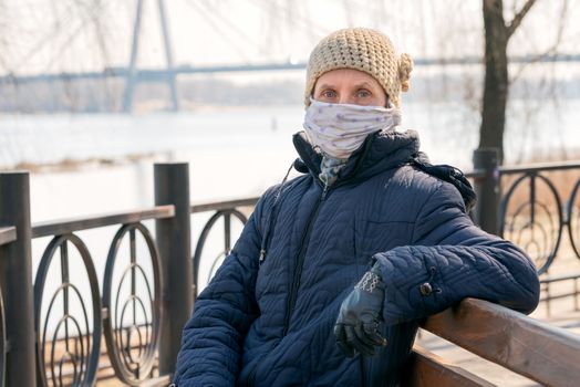 A poor elderly woman wears a homemade mask to protect herself from viruses such as coronavirus, also known as covid-19, or SARS and MERS. She sits on a bench close to the river