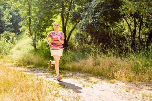 A happy senior woman is running with the sun in backlight, toward the shadow, in the forest during a warm summer day