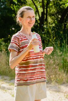 A senior woman is drinking water or yogurth to quench her thirst after a long run in the forest