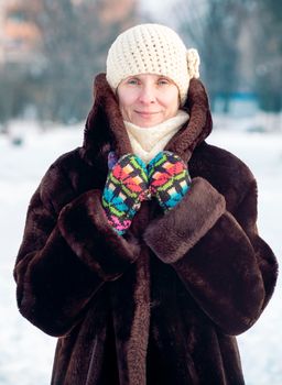 A winter portrait of a smiling senior adult woman wearing a wool cap, a scarf and colored gloves, with a snow background