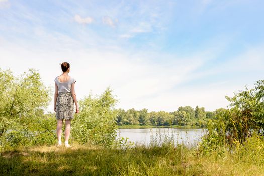 A woman standing up close to the Dnieper river in Kiev, Ukraine, observes in the distance from a raised point of view, under a soft cloudy summer sky