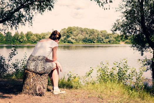 A sad and nostalgic woman is sitting close to the Dnieper river in Kiev, Ukraine. She is thinking while watching or observing far in the distance, under a warm and soft summer sun