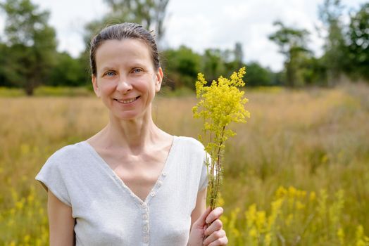 Portrait of an adult woman standing up in a meadow covered with Galium verum flowers, also known as lady's bedstraw or yellow bedstraw, with a bunch of yellow flowers in her hands, under the warm and soft summer sun