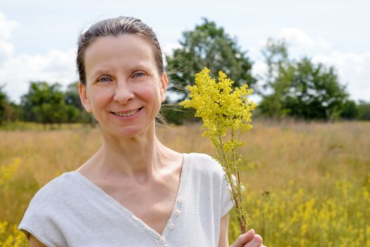 Portrait of an adult woman standing up in a meadow covered with Galium verum flowers, also known as lady's bedstraw or yellow bedstraw, with a bunch of yellow flowers in her hands, under the warm and soft summer sun