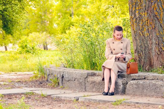 Elegant businesswoman sitting on a stone wall during a sunny spring day, and looking a message or dialing a number on her mobile phone