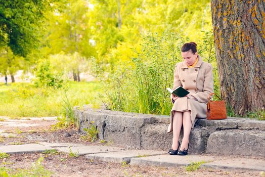 Elegant businesswoman sitting on a stone wall during a sunny spring day, and reading an adventure book