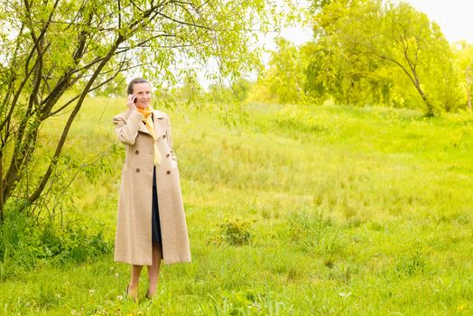 An elegant senior businesswoman, with a coat and a yellow scarf, calling with her mobile phone in the park under the spring sun