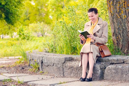 Elegant businesswoman sitting on a stone wall during a sunny spring day, and reading a funny book